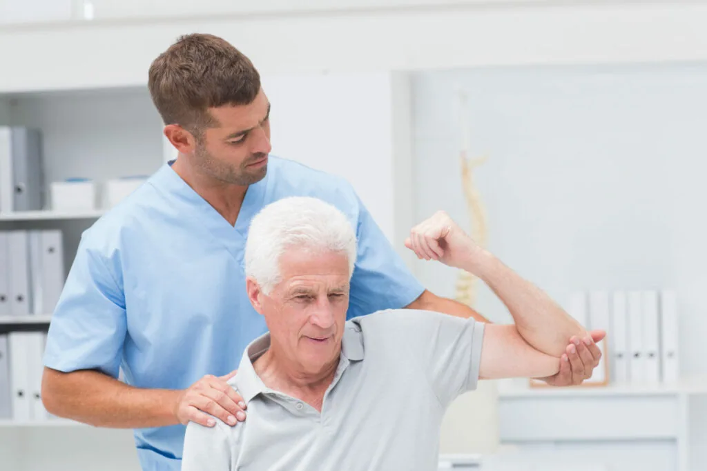 Healthcare provider in blue scrubs performing shoulder examination on elderly male patient in clinical setting