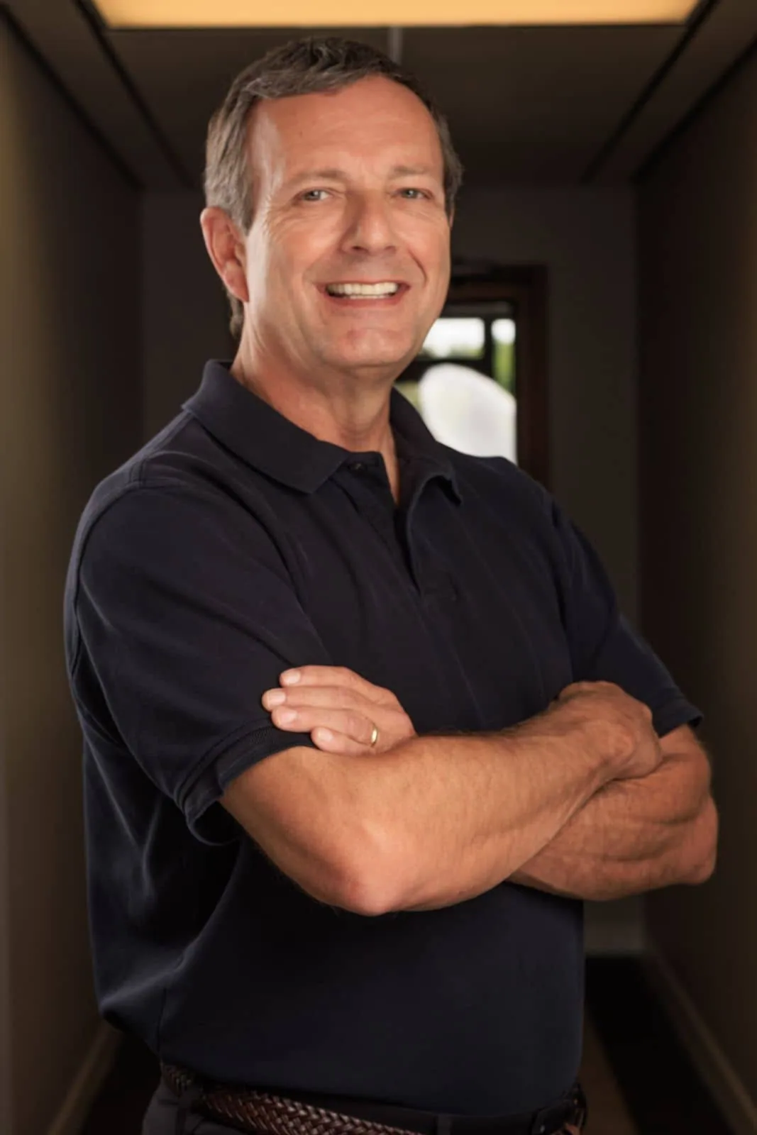 Middle-aged man with gray hair smiling at camera, arms crossed, wearing dark blue polo shirt in hallway